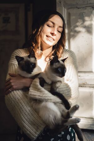 Attractive girl embracing her Siamese cats. Indoor portrait of cute woman playing with her two pets.の写真素材