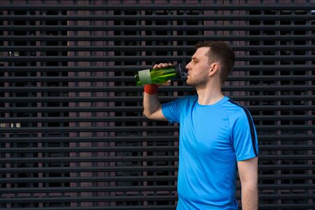 Profile view of sporty young man drinking water from bottle after long city run on urban gray backgroundの写真素材