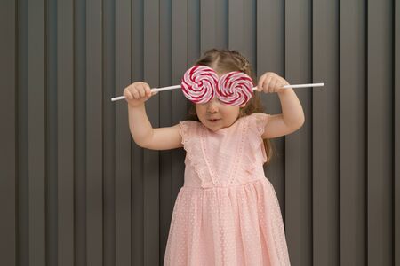Happy young caucasian little girl kid with happy big sweet lollypops candy over her eyes on gray striped backgroundの写真素材