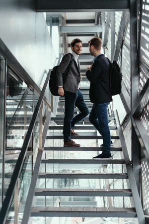 Two young businessmen going up, down the stairs while chatting in a modern building.の写真素材