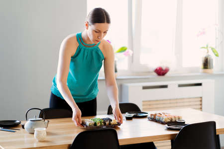 Young woman waiting for guests, friends while arranging the table with various sushi sets in a sunny home with window in the backgroundの写真素材