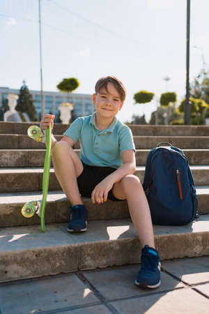 School Boy in blue polo shirt sitting on the stairs with a blue backpack and green penny board