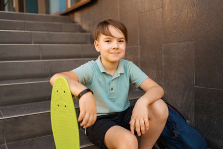 School Boy in blue polo shirt sitting on the stairs with a blue backpack and green penny boardの写真素材