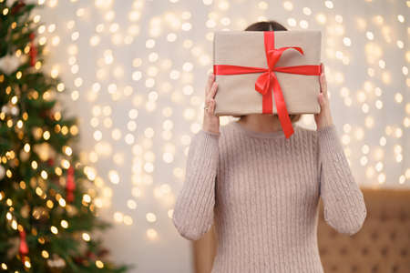 Portrait of young happy woman red lips hiding behind wrapped gift box. Close up satisfied woman received present box. Festive Christmas lights background.の写真素材