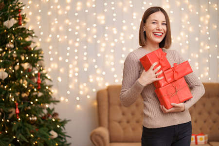 Happy young woman holding many present boxes with Christmas tree and lights in backgroundの写真素材