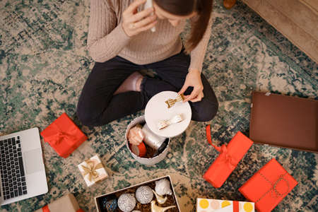 Woman wrapping Christmas gift and talking on the phone while sitting in Christmas decorated living