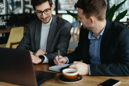 Two businessmen talking about new opportunities sitting with laptop at desk, planning project, considering business offerの写真素材
