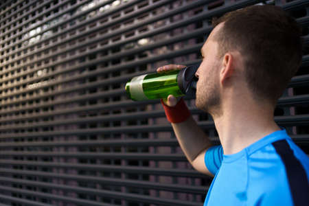 Profile view of sporty young man drinking water from bottle after long city run on urban gray backgroundの写真素材