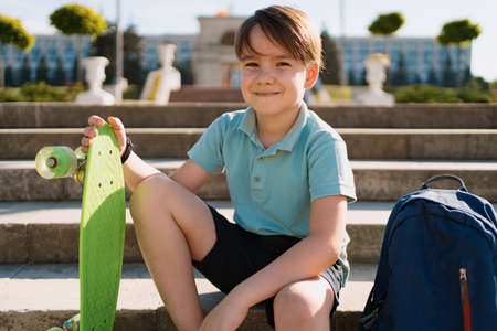 School Boy in blue polo shirt sitting on the stairs with a blue backpack and green penny boardの写真素材