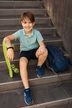 School Boy in blue polo shirt sitting on the stairs with a blue backpack and green penny boardの写真素材