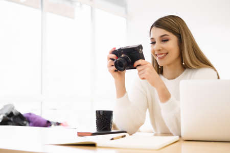 Portrait of beautiful photographer woman working in office holding camera with laptop. Bloger...