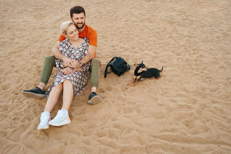 Beautiful couple relaxing on sand hugging and kissing while playing with their positive dogの写真素材