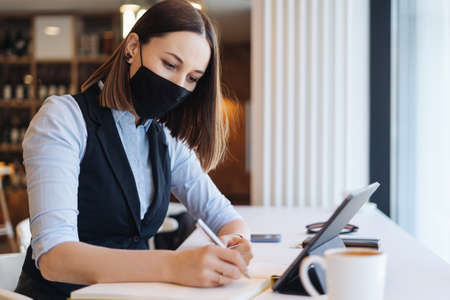 Pretty woman wearing mask planning working schedule writing in notebook while sitting at working...
