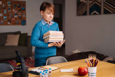 Distance online education, internet learning. First grade boy studying at home, holding a bunch of books, getting ready for online lessonの写真素材