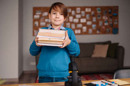 Distance online education, internet learning. First grade boy studying at home, holding a bunch of books, getting ready for online lessonの写真素材