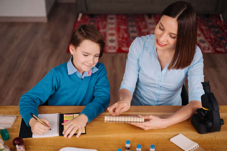 Aunt and nephew do homework together. Women explain kid writes in notebook. Schoolboy studying at home. Family relationships, primary education, childhoodの写真素材