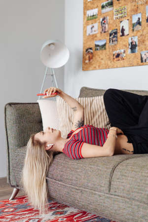 Woman listening music in headphones while lying on sofa in living room looking to smartphone screen