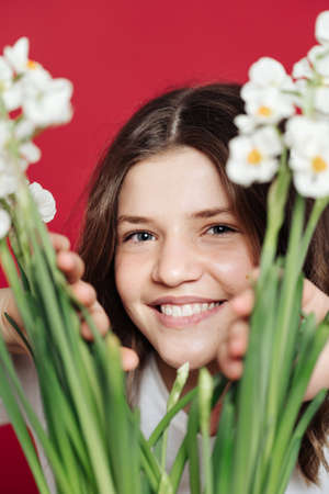 Close up portrait of a smiling teenager girl looking among blooming flowers over red background