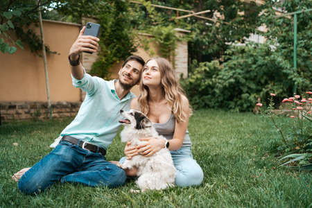 Beautiful happy couple making selfie with their lovely dog on the backyard while sitting on the grassの写真素材