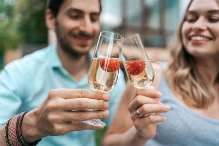 Happy couple portrait clinking two glasses with sparkling wine and strawberries inside with blurred house on background. Celebrating loveの写真素材
