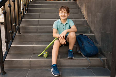 School Boy in blue polo shirt sitting on the stairs with a blue backpack and green penny boardの写真素材
