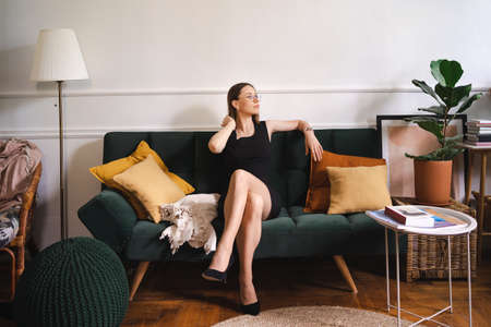 Confident businesswoman wearing glasses in a black classic dress sitting in a modern apartment on a couch. Green plush sofa with yellow pillowsの写真素材