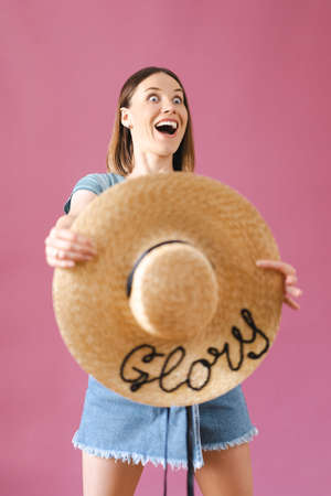 Portrait of joyful young woman in casual wearing straw hat on pink background. Cheerful young woman...