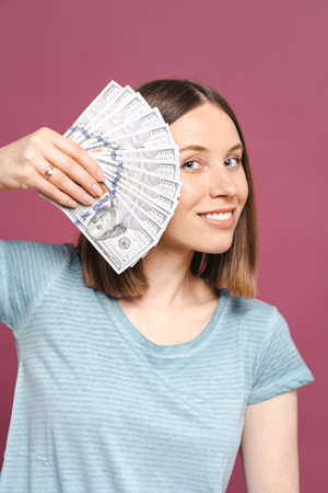 Funny young lady in a blue summer tshirt is holding money and looking to them over a pink background...