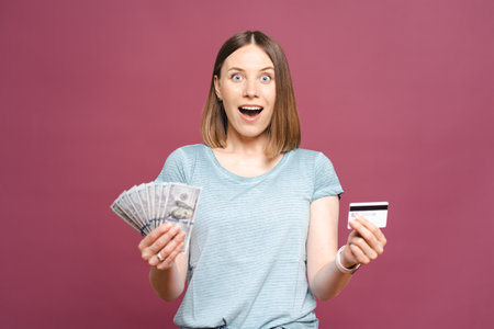 Close up portrait of a smiling woman in blue tshirt holding a credit card in one hand and cash in thensecond isolated on pink backgroundの写真素材