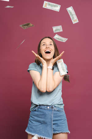 Photo of a surprised young girl reacting excited to flying money over a pink background, making a winner gesture.の写真素材