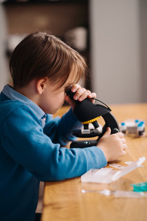 Distance online education, internet learning. First grade boy studying at home using microscope, making notes, biology online lessonの写真素材