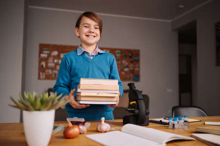 Distance online education, internet learning. First grade boy studying at home, holding a bunch of books, getting ready for online lessonの写真素材