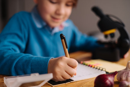 Distance online education, internet learning. First grade boy studying at home using microscope, making notes, biology online lessonの写真素材