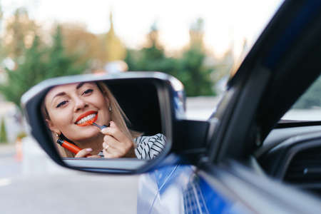 Young Caucasian woman applying lipstick looking at reflection in car mirror.の写真素材