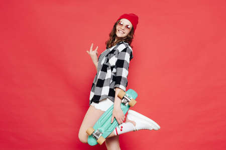 Happy stylish girl teenager wearing glasses, red beanie, white shorts and plaid shirt posing with...