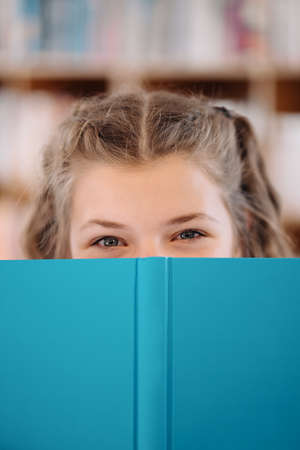 Portrait of a pretty young girl hiding behind an open blue book and looking to camera over bookshelf backgroundの写真素材