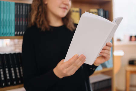 Teen girl among a pile of books. A young girl reads a book with shelves in the background. She is...