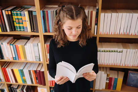 Teen girl among a pile of books. A young girl reads a book with shelves in the background. She is surrounded by stacks of books. Book day.の写真素材