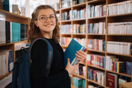 Happy teenage girl or student wearing glasses taking book from shelf in library, wear backpack- People, knowledge, education and school conceptの写真素材