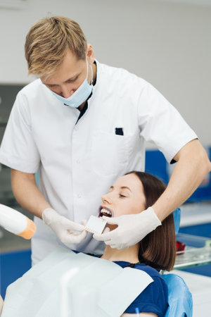 Close up portrait of beautiful young lady sitting in dental chair while stomatologist hands in sterile gloves holding tooth samples. She is smilingの写真素材