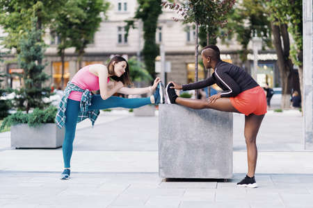 Two fit diverse young woman working out together in city square doing stretching exercise and hand extensionsの写真素材