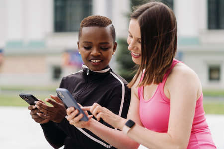 Cheerful smiling friends in sportswear sitting on a bench in the city discussing while using...