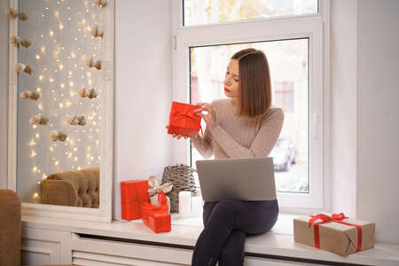 Smiling young woman greeting friends with Christmas in video chat on laptop with gift boxes surrounding her near window. Female order sitting on window.の写真素材