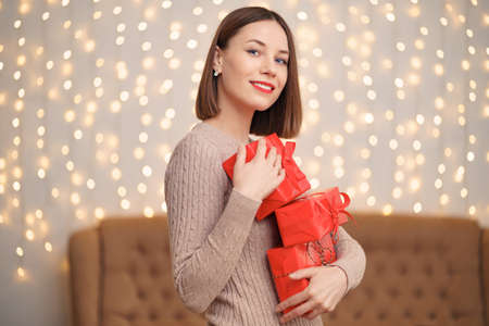 Happy young woman holding many present boxes with Christmas tree and lights in backgroundの写真素材