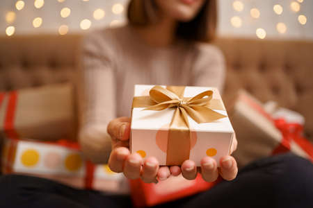 Happy young woman holding a present showing surrounded by gift boxes sitting crossed legs on a camel sofa with lights in backgroundの写真素材