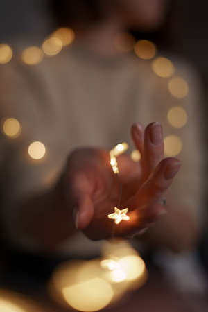 Woman holds a glowing star in her hand to make a wish. Lady hand hold Christmas decoration with bokeh lightsの写真素材