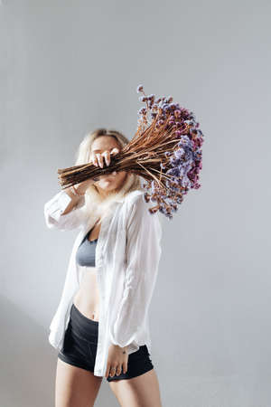 Portrait of a beautiful young girl in white shirt, black top and shorts, holding a big bouquet of dried flowers over gray background splashed by lines of lightの写真素材