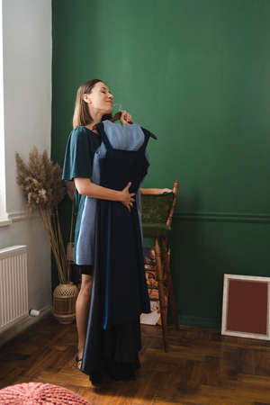 Charming dark blond caucasian woman in green dress smiles widely and holds hanger with dresses dress, trying to decide what to wear.の写真素材