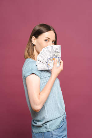 Funny young lady in a blue summer tshirt is holding money and looking to them over a pink background...