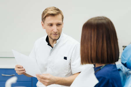 Male Professional Dentist With Gloves And Mask Holding papers photo And Show What The Treatment Will Look Like Of The Patients Teeth. Discussion Of The Treatment Plan And Healthy Smileの写真素材